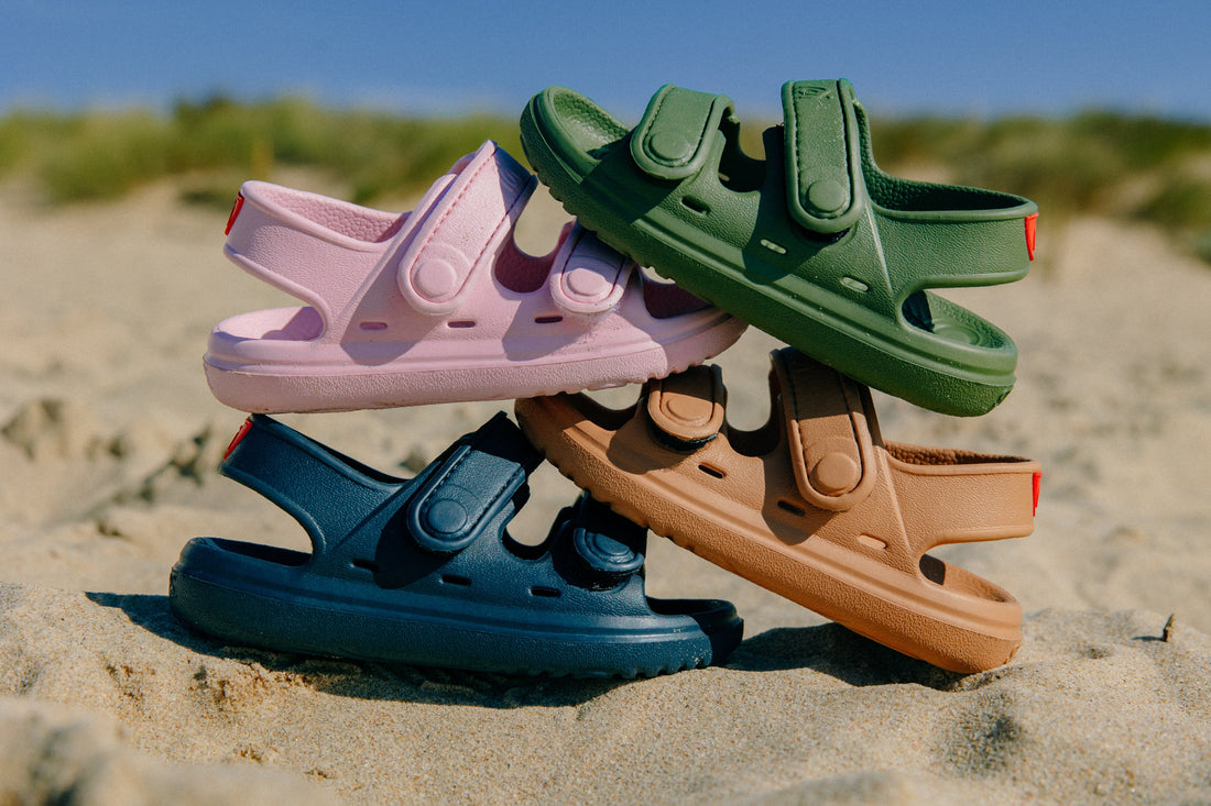 A stack of Wonderskips sandals in pink, olive green, navy blue, and tan, showing the full range of colors on a beach.