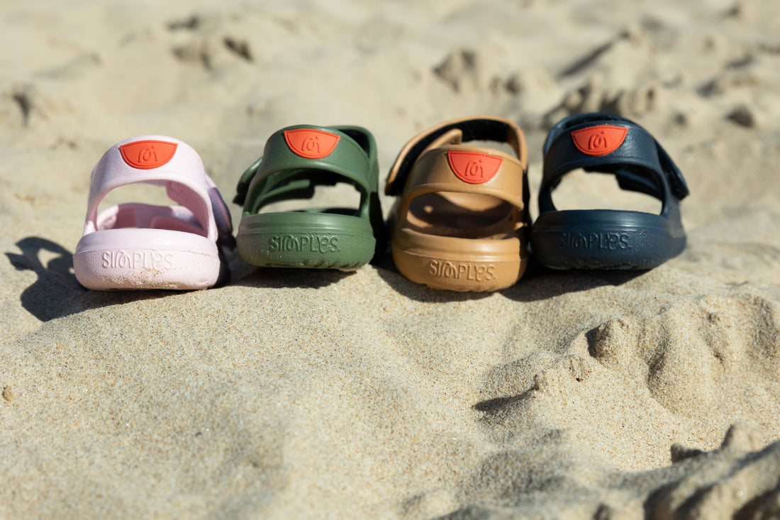 Row of Wonderskips toddler sandals in pink, olive green, tan, and navy blue sitting on beach sand.