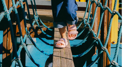 Toddler wearing pink Wonderskip sandals walking across a rope bridge at a playground, balancing on a wooden beam.