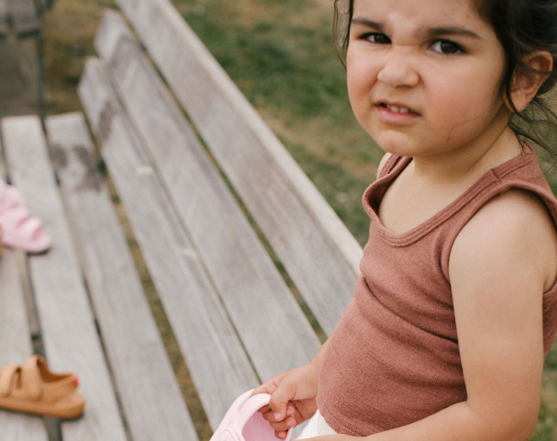 A child holding a pink Wonderskips sandal while sitting on a wooden bench, with additional tan Wonderskips foam sandals visible in the background.